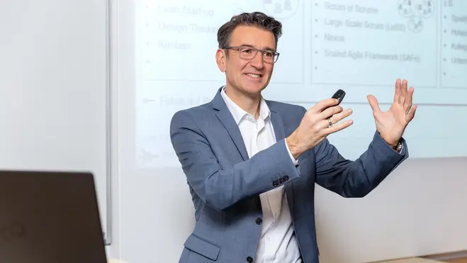 Ein Mann in einem grauen Blazer hält einen Präsentationsclicker und spricht enthusiastisch vor einer Projektionswand. Im Hintergrund sind verschiedene Diagramme und Begriffe zu sehen, die mit agilen Methoden und Frameworks in Verbindung stehen.