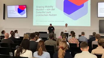 Eine Präsentation zum Thema "Sharing Mobility Monitor – von der Datenflut zum praktischen Nutzen" wird gehalten. Zwei Referenten stehen vorne vor einem Publikum, das aufmerksam zuhört. Im Hintergrund sind Bildschirme mit den Präsentationsinhalten zu sehen.