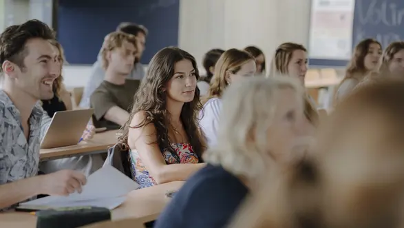 Man sieht einen Raum voller Studierenden, die gespannt nach vorne schauen und dem Kurs zuhören. Sie sitzen an Schreibtischen und haben ihre Tablets vor sich. Im Hintergrund sieht man Tafelen.