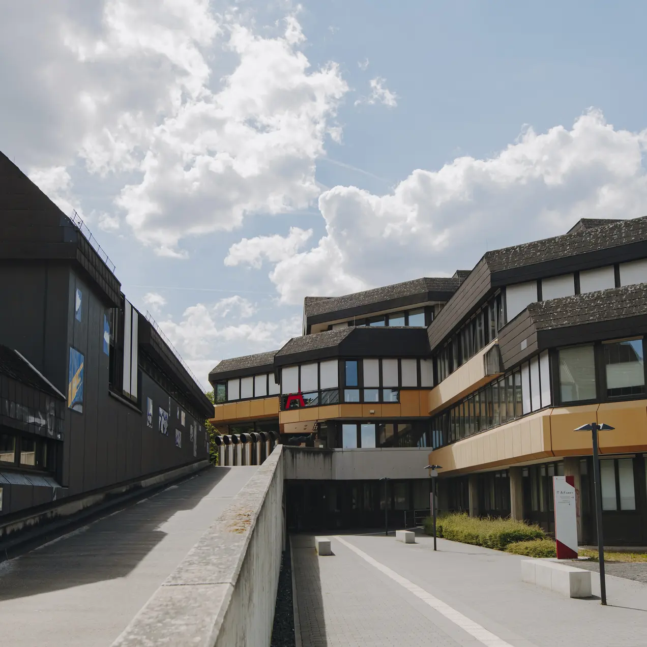 Blick auf ein modernes Gebäudeensemble mit mehreren Etagen. Die Architektur ist funktional und hat eine Kombination aus dunklen und hellen Fassaden. Im Vordergrund verläuft ein gepflasterter Weg, auf dem Fahrräder abgestellt sind. Helle Wolken sind am Himmel sichtbar.