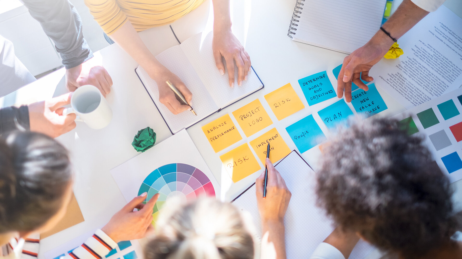 Workshop Gruppenarbeit Verschiedene Post-Its auf einem Tisch, um den mehrere Menschen rundherum stehen. Einige zeigen auf Post-Its