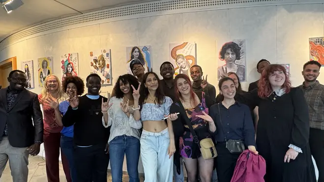 Eine Gruppe von 15 Personen steht zusammen in einem Raum und posiert für ein Foto. Im Hintergrund hängen verschiedene Kunstwerke an der Wand. Die Personen lachen und zeigen teils Friedenszeichen oder andere Gesten.