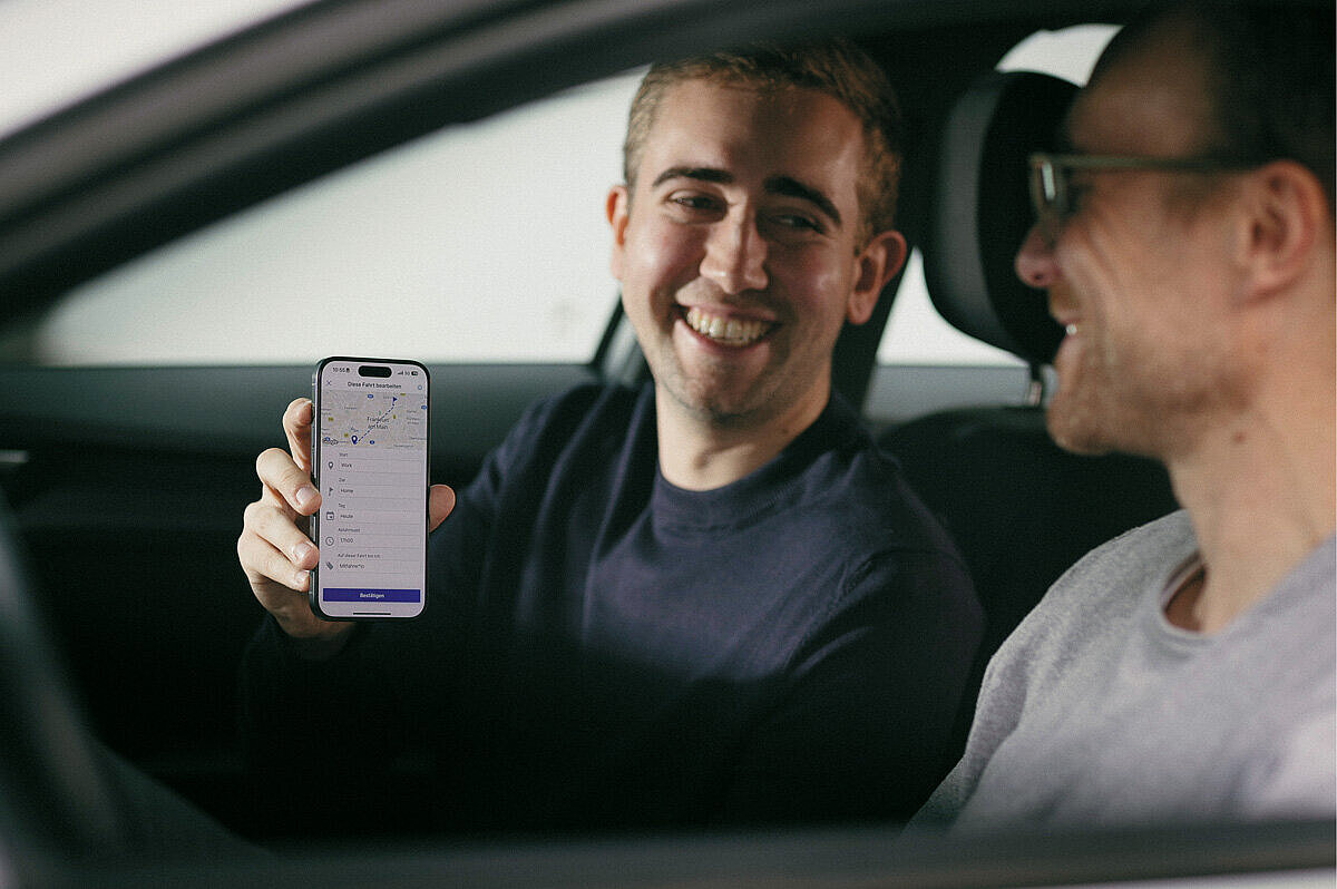 Zwei Männer sitzen in einem Auto und lächeln sich an, einer hält ein Smartphone in der Hand