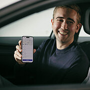 Zwei Männer sitzen in einem Auto und lächeln sich an, einer hält ein Smartphone in der Hand