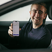 Zwei Männer sitzen in einem Auto und lächeln sich an, einer hält ein Smartphone in der Hand