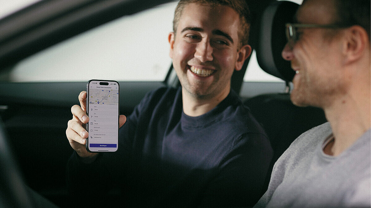 Zwei Männer sitzen in einem Auto und lächeln sich an, einer hält ein Smartphone in der Hand