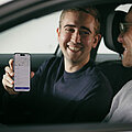 Nutzer der neuen Mitfahrplattform der Fraport AG Zwei Männer sitzen in einem Auto und lächeln sich an, einer hält ein Smartphone in der Hand