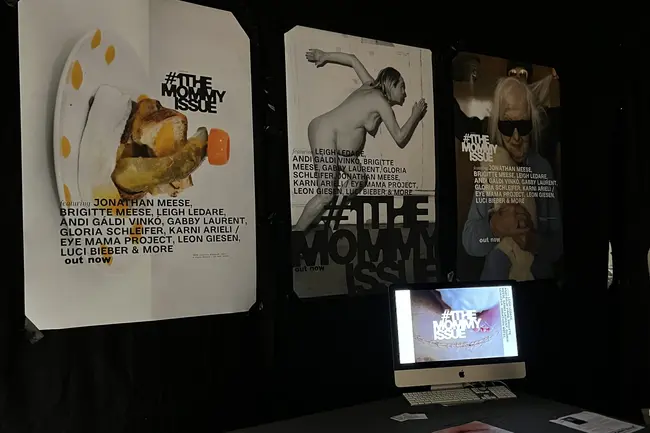 Eine Ausstellung mit drei Posters, die das Thema "The Mommy Issue" präsentieren. Im Vordergrund ein Tisch mit einem Computer und Druckmaterialien. Die Atmosphäre ist dunkel, um die Kunstwerke hervorzuheben.