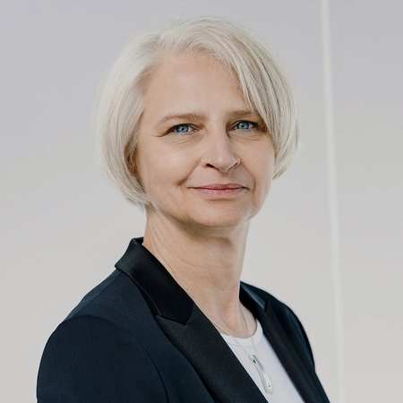 Porträt einer Frau mittleren Alters mit kurzen, blonden Haaren und blauen Augen. Sie trägt einen schwarzen Blazer über einem weißen Oberteil und lächelt freundlich. Der Hintergrund ist neutral, wodurch der Fokus auf ihrem Gesicht liegt.