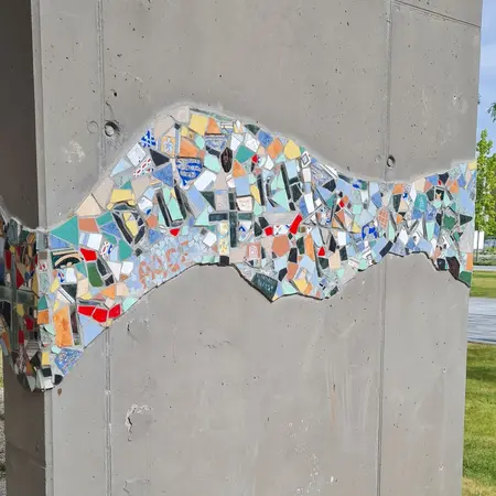 Das Bild zeigt ein Mosaik aus bunten Keramikscherben, das auf einer Betonsäule angebracht ist. Die Form des Mosaiks erinnert an eine Welle oder einen Fluss. Die Farben variieren zwischen Blau, Grün, Rot und Gelb, wodurch ein lebhaftes und kreatives Kunstwerk entsteht.