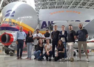 Eine Gruppe von sieben Personen steht vor einem großen Flugzeug, der Super Guppy. Hinter ihnen sind Teile des Flugzeugs deutlich sichtbar. Die Personen haben unterschiedliche Ausdrucksweisen und tragen legere Kleidung. Die Szene spielt in einer großen Halle.
