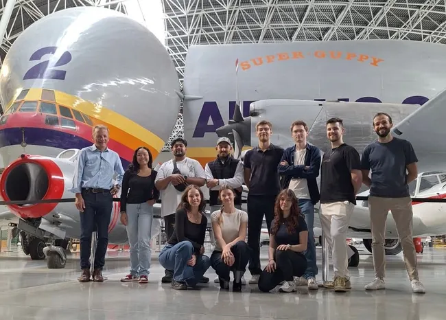 Eine Gruppe von sieben Personen steht vor einem großen Flugzeug, der Super Guppy. Hinter ihnen sind Teile des Flugzeugs deutlich sichtbar. Die Personen haben unterschiedliche Ausdrucksweisen und tragen legere Kleidung. Die Szene spielt in einer großen Halle.