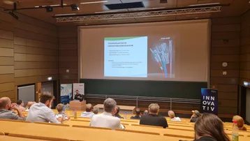 Ein Referent steht vor einer Präsentation in einem Seminarraum. Die Präsentation zeigt Informationen zu Hochgeschwindigkeitslösungen mit einem Bild von Kabeln. Im Publikum sitzen Teilnehmer, die dem Vortrag aufmerksam folgen. Rechts steht ein Banner mit dem Wort "INNOVATIV".
