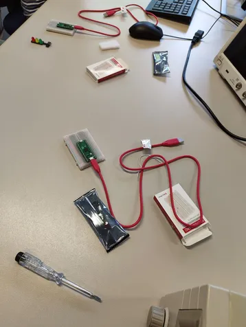 Auf dem Tisch liegen verschiedene elektronische Komponenten und Werkzeuge. Im Vordergrund befindet sich ein Bauteil mit roten Kabeln, das mit einem USB-Anschluss verbunden ist. Daneben sind Verpackungen und ein Schraubendreher zu sehen. Ein Computer und ein Monitor sind im Hintergrund.