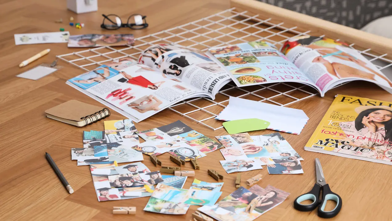 Auf einem Tisch liegen verschiedene Zeitschriften, Fotos und ein Notizbuch. Scissors und Büroklammern sind ebenfalls sichtbar. Die Anordnung deutet auf kreative Arbeit hin, möglicherweise für ein Vision Board oder ein Scrapbook. Der Tisch hat eine helle Holzoberfläche.