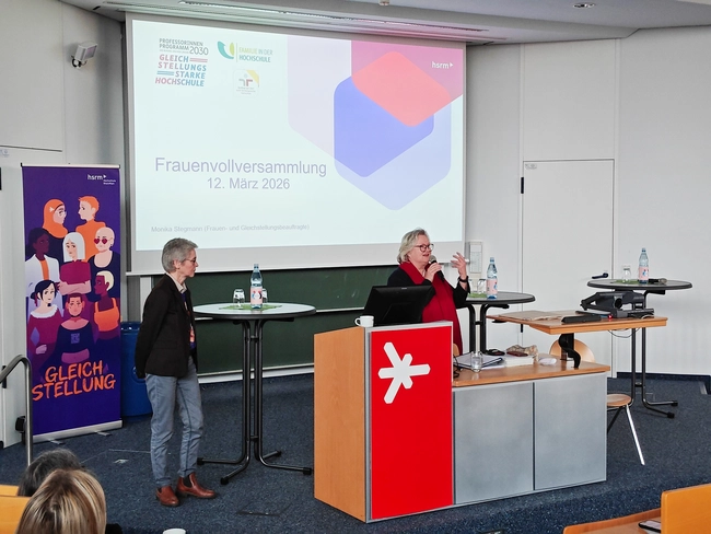 Auf der Frauenvollversammlung am 12. März 2026 stehen zwei Frauen an Podien. Eine redet und gestikuliert, während die andere aufmerksam zuhört. Im Hintergrund ist ein Banner mit dem Titel "Gleichstellung" zu sehen, das das Thema der Veranstaltung unterstreicht.