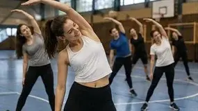 Eine Gruppe junger Menschen übt in einer Sporthalle Dehnübungen. Die Vordergrundperson, eine Frau in einem weißen Top, beugt sich zur Seite. Die anderen Teilnehmer folgen synchron, alle tragen sportliche Kleidung. Die Atmosphäre vermittelt Energie und Aktivität.
