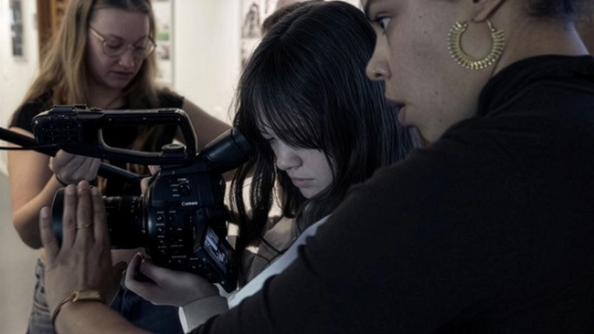 Drei Personen befinden sich in einem Raum, während sie eine Kamera bedienen. Eine Frau hält die Kamera und konzentriert sich darauf, während eine andere Frau ihr Anweisungen gibt. Eine dritte Person beobachtet diesen Prozess. Der Fokus liegt auf dem gemeinsamen Lernen zur Filmproduktion.