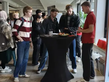 Eine Gruppe von vier Personen steht an einem Stehtisch in einem Innenbereich. Zwei junge Männer unterhalten sich mit einem Mann, der ein rotes Shirt trägt. Auf dem Tisch liegen Unterlagen und Snacks, während andere Besucher im Hintergrund warten.