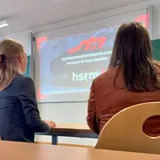 Zwei Personen sitzen an einem Tisch und schauen auf eine Projektionsleinwand. Auf der Leinwand sind die Worte "UNTERNEHMENSVORSTELLUNG - Personal & Organisation" und "hsrm" zu sehen. Der Hintergrund ist unscharf und es scheint sich um eine Präsentation zu handeln.