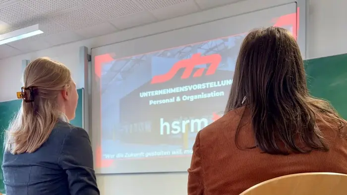 Zwei Personen sitzen an einem Tisch und schauen auf eine Projektionsleinwand. Auf der Leinwand sind die Worte "UNTERNEHMENSVORSTELLUNG - Personal & Organisation" und "hsrm" zu sehen. Der Hintergrund ist unscharf und es scheint sich um eine Präsentation zu handeln.