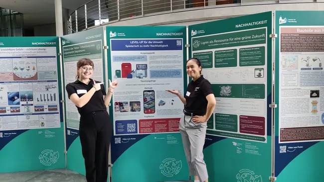 Zwei Personen stehen vor einem Ausstellungsposter zu Nachhaltigkeit. Die Frau auf der linken Seite zeigt auf ein Plakat, während der Mann auf der rechten Seite ebenfalls auf die Poster verweist. Die Poster enthalten Informationen zu nachhaltigen Lösungen und Projekten.