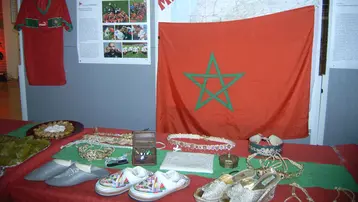 Ausstellung über Marokko mit einer roten Flagge, die einen grünen Stern zeigt, einer Karte des Landes, einem traditionellen marokkanischen Kleidungsstück, und verschiedenen handgefertigten Gegenständen wie Schuhen und Schmuck, die die Kultur und das Erbe Marokkos repräsentieren.