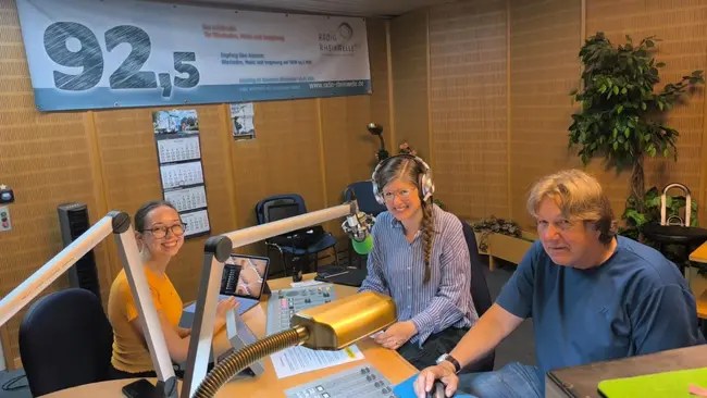 Drei Personen sitzen an einem Radiotisch. Eine Frau mit Kopfhörern lächelt in die Kamera, während ein Mann und eine weitere Frau ebenfalls am Tisch sitzen. Der Hintergrund zeigt eine Radiostation mit einem Banner und Steuerpult.