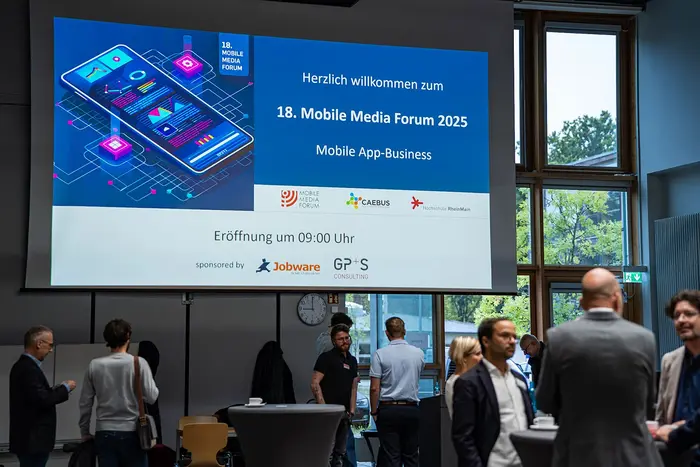 Teilnehmer:innen des Mobile Media Forums 2025 stehen unter einer großen Leinwand, auf der das Key Visual der Veranstaltung gezeigt wird, und unterhalten sich.