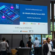 Teilnehmer:innen des Mobile Media Forums 2025 stehen unter einer großen Leinwand, auf der das Key Visual der Veranstaltung gezeigt wird, und unterhalten sich.