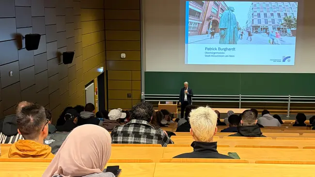 Ein Vortrag von Patrick Burghardt findet in einem Hörsaal statt, während eine Präsentation im Hintergrund angezeigt wird. Die Anwesenden sitzen in Reihen und hören aufmerksam zu.