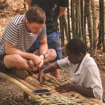 Three students on an excursion in Ghana are in a natural bamboo environment. A woman demonstrates how to weave natural materials, while the two men stand behind her and watch intently.
