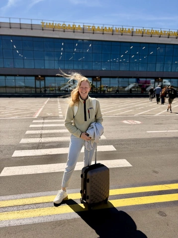 Eine junge Frau mit langen, blonden Haaren steht auf einem Zebrastreifen vor einem großen Gebäude mit Glaselementen. Sie trägt einen hellen Pullover und hält einen Koffer in der Hand. Die Szenerie ist sonnig und zeigt einen internationalen Flughafen im Hintergrund.