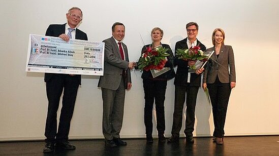 Menschen stehen nebeneinander. Ein Mann hält eine große Scheck-Tafel in der Hand und zwei weitere Personen haben einen Strauß Blumen in der Hand. 