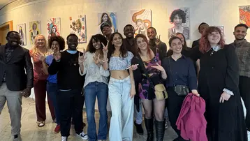 Eine Gruppe von 15 Personen steht zusammen in einem Raum und posiert für ein Foto. Im Hintergrund hängen verschiedene Kunstwerke an der Wand. Die Personen lachen und zeigen teils Friedenszeichen oder andere Gesten.