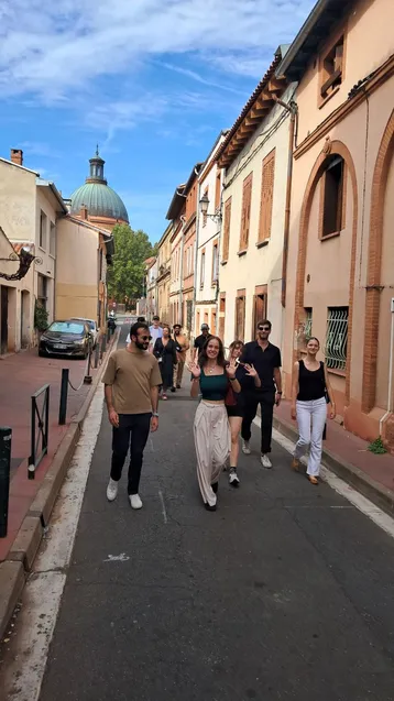 Eine Gruppe von sechs Personen geht fröhlich eine belebte Straße entlang. Die Gebäude sind in warmen Farben gehalten, und im Hintergrund ist eine Kuppel zu sehen. Die Atmosphäre wirkt entspannt und gesellig.