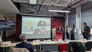 A presentation in a conference room, with several people standing at a podium. In the background, there is a screen with illustrations and texts promoting creativity and collaboration. Some audience members are seated at tables watching the presentation.