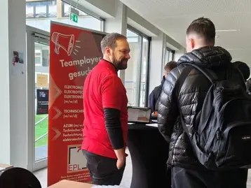 Ein Mann in einem roten T-Shirt steht an einem Informationsstand und spricht mit einer Person im schwarzen Outfit. Im Hintergrund ist ein Banner mit der Aufschrift "Teamplayer gesucht" sowie ein Tisch mit Snacks und Informationsmaterialien sichtbar.