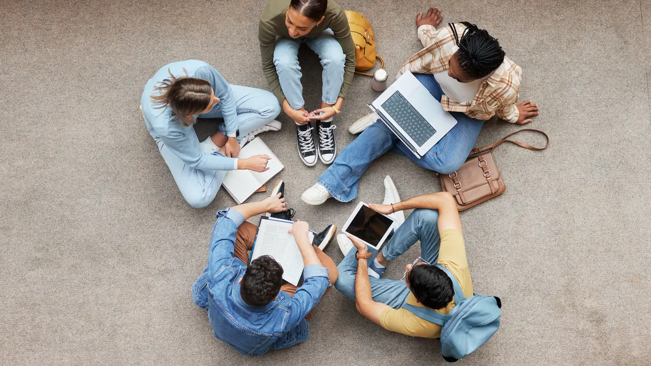 Sechs junge Erwachsene sitzen im Kreis auf dem Boden. Sie arbeiten gemeinsam, nutzen Laptops, Notizbücher und Tablets. Einige schreiben, während andere diskutieren. Die Atmosphäre wirkt kooperativ und konzentriert.