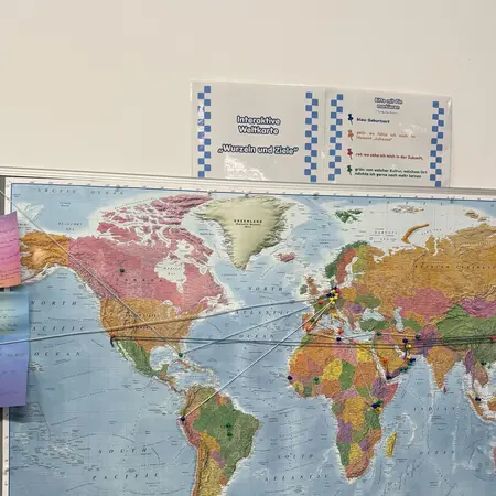 Eine Weltkarte hängt an der Wand. Sie zeigt die Kontinente und Länder der Erde in verschiedenen Farben. Einige Zettel sind an die Karte geheftet. Unterhalb der Karte sind Flaggen verschiedener Länder abgebildet. Oben steht eine Tafel mit Informationen oder Anweisungen.
