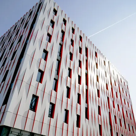 Das Bild zeigt eine moderne Architektur eines Gebäudes mit einer auffälligen Fassade aus grauen und roten Paneelen. Die Sicht ist von unten nach oben gerichtet, wobei die Fenster der oberen Etagen sichtbar sind. Der klare Himmel im Hintergrund unterstreicht die elegante Form des Gebäudes.