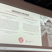 Auf einer Präsentationsfolie ist ein Diplom zu sehen mit einem Foto eines jungen Mannes, seinem Namen und Alters sowie einer kurzen Biografie. 