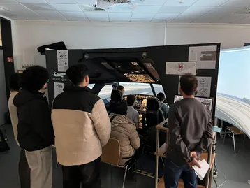 Eine Gruppe von Menschen beobachtet einen Flugsimulator. Im Inneren des Simulators befinden sich Steuerungen und Bildschirme, die eine virtuelle Flugumgebung anzeigen. Die Teilnehmer scheinen interessiert zu sein und stehen in einer Reihe hinter dem Simulator.