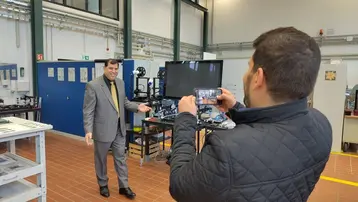 Ein Mann in einem Anzug steht in einem modernen Raum mit technischen Geräten und blickt zur Kamera. Ein weiterer Mann hält ein Smartphone und fotografiert ihn. Im Hintergrund sind Regale und Arbeitsstationen sichtbar.