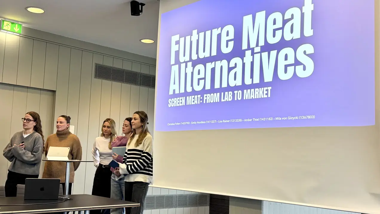 Eine Gruppe von fünf Frauen steht vor einer großen Präsentationsleinwand, auf der der Titel „Future Meat Alternatives: Screen Meat - From Lab to Market“ zu sehen ist. Sie diskutieren über alternative Fleischprodukte aus dem Labor. Ein Laptop ist auf dem Tisch vor ihnen platziert.