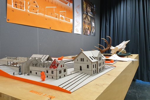 Ausstellungspräsentation der „Akadamie für Jagdkultur und Natur“ von Lilia Dalmann. Auf dem Tisch liegen einige für die Jagd typische Exponate, an der Wand dahinter sind die Raumkonzepte zu sehen. Im Vordergrund steht ein Miniaturmodell der Gebäude.