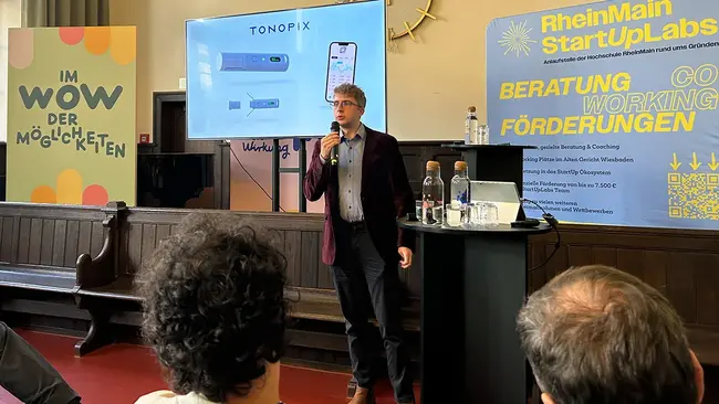 Marco Großmann, Student der Hochschule RheinMain, stellt seine Gründungsidee vor. Mit einem Mirkofon in der Hand spricht er zum Publikum im Heimathafen im Alten Gericht.