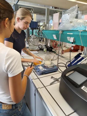Zwei Frauen stehen in einem Labor und arbeiten an einem Experiment. Eine von ihnen wiegt eine Substanz auf einer digitalen Waage, während die andere sie mit einem Instrument unterstützt. Im Hintergrund sind verschiedene Laborgeräte sichtbar.