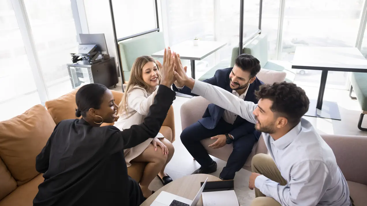 Eine Gruppe von vier Personen in einem modernen Büro feiert gemeinsam einen Erfolg. Sie geben sich gegenseitig High-Fives und wirken glücklich und motiviert. Der Fokus liegt auf der positiven Teamdynamik und Zusammenarbeit. Ein Laptop und Notizen sind auf dem Tisch sichtbar.