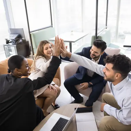 Eine Gruppe von vier Personen in einem modernen Büro feiert gemeinsam einen Erfolg. Sie geben sich gegenseitig High-Fives und wirken glücklich und motiviert. Der Fokus liegt auf der positiven Teamdynamik und Zusammenarbeit. Ein Laptop und Notizen sind auf dem Tisch sichtbar.